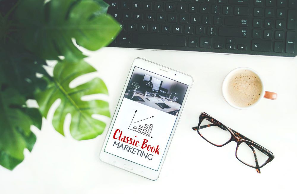 Image of laptop with a plant leaf in top left corner. On the desk is a tablet with the words "online book marketing" and a graph. A pair of glasses are to the right of the tablet and a cup of coffee is above them.