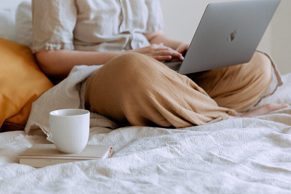 A person sits on their bed with their computer on their lap.