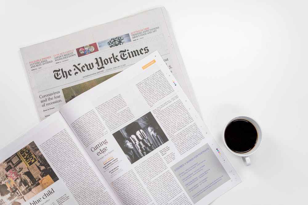 A copy of the New York Times newspaper sits open and horizontal on a white table with a cup of black coffee next to it.