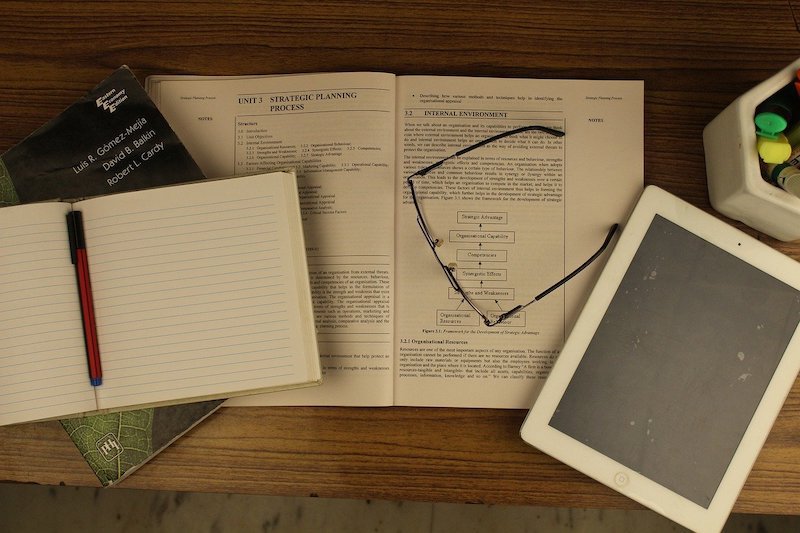 aerial shot of a wooden desk with an open book, notebook, ereader, and pair of glasses.