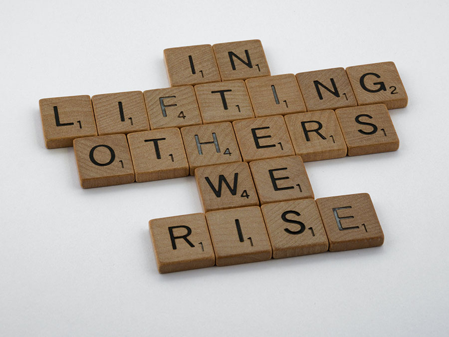 scrabble pieces spelling "IN LIFTING OTHERS WE RISE"