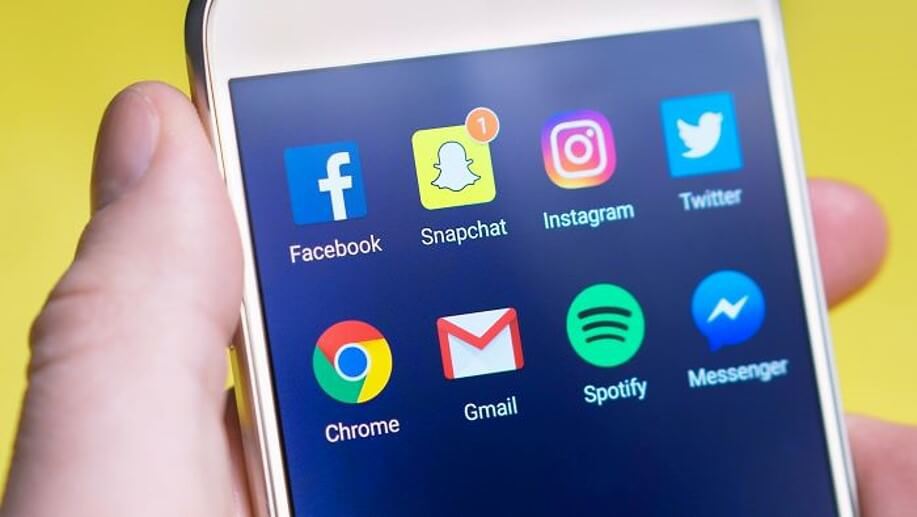 Hand holding a mobile phone showing two rows of commonly used apps. First row: Facebook, Snapchat, Instagram, and Twitter. Second row: Chrome, Gmail, Spotify, and Messenger.