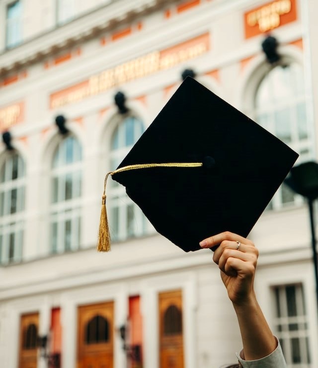 a hand holding a gold-tasseled, black mortarboard graduation hat in front of a university building