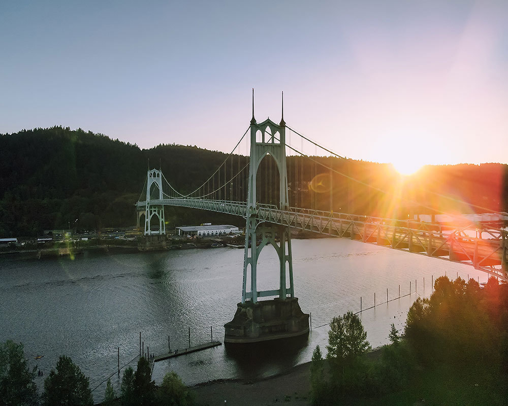 The sun setting over the St. John's Bridge, a large, blue-green suspension bridge.