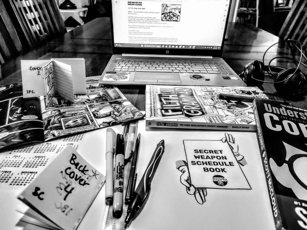 black and white photo of a desk with laptop, pens, comic images on paper, books about comics editing