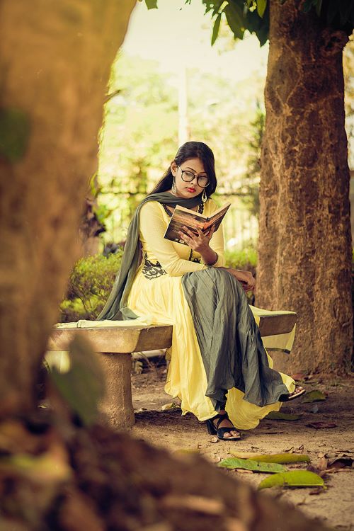 South Asian woman in a green and yellow dress reading a book with legs crossed on a bench outside between two trees