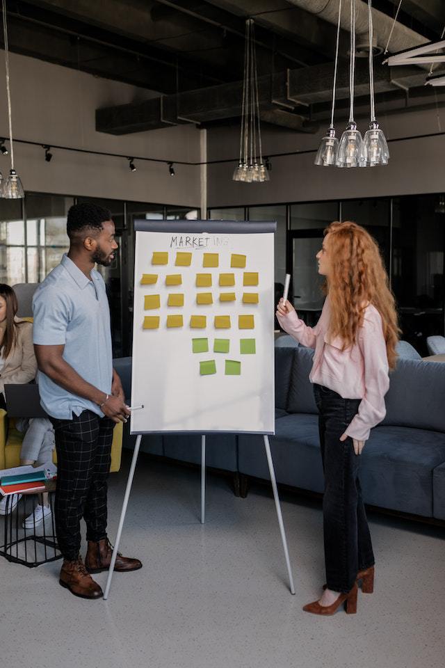 A man and woman standing on either side of a stand-up board filled with sticky notes.